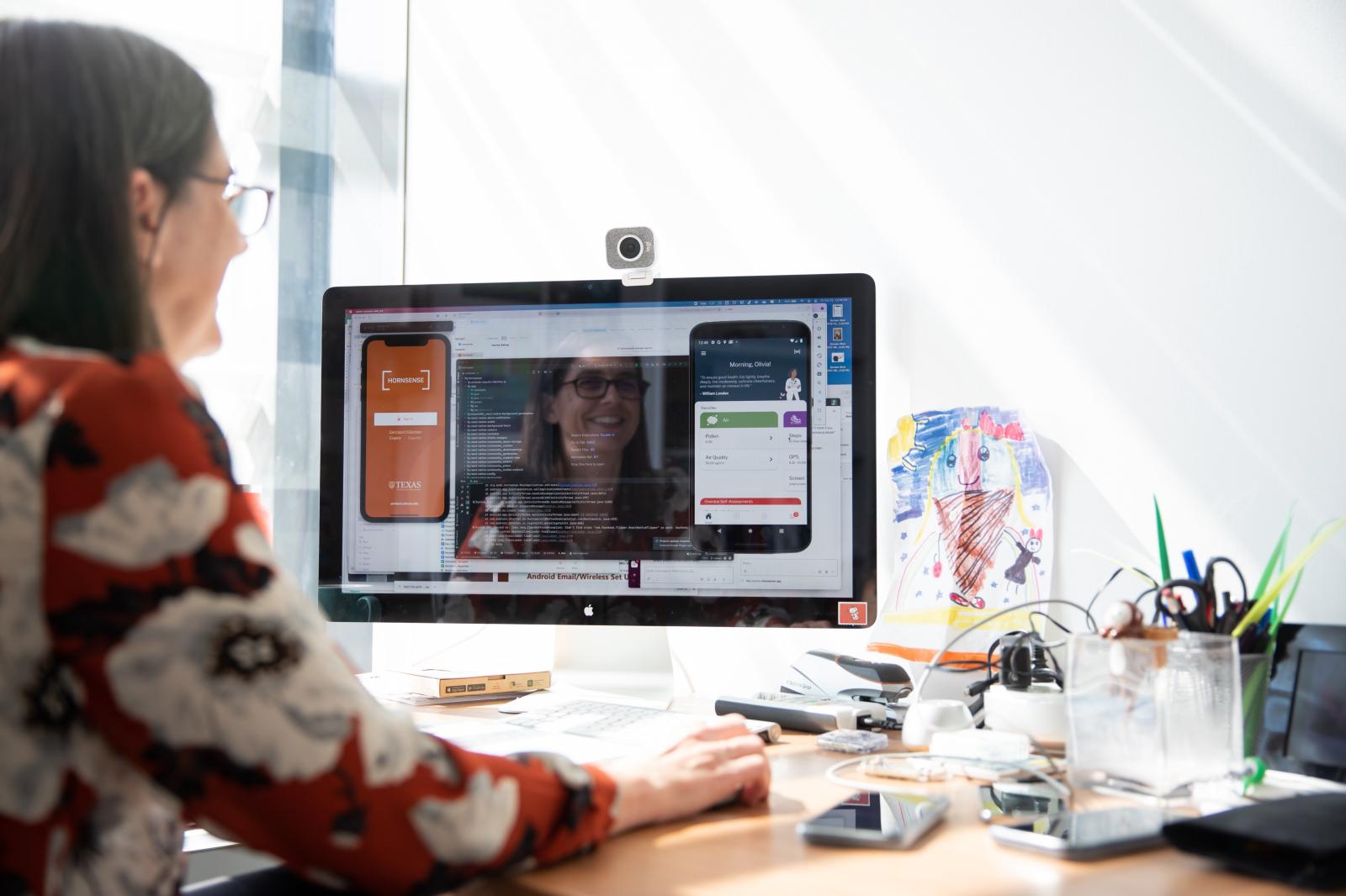 A woman sits at her desktop computer in her brightly-lit office designing a mobile application.