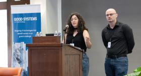 Project co-leaders Azza El-Masri, a graduate student, and Brad Limov, a postdoctoral researcher, present their findings at the 2025 Good Systems Symposium at UT Austin. El-Masri speaks behind a lectern; Limov stands next to her. Behind them is a Good Systems-branded roll-up banner. 