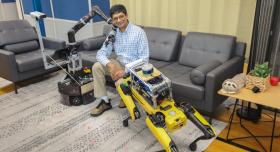 Joydeep Biswas, an associate professor of computer science who will lead the team awarded the NSF grant, sits on a couch smiling, flanked by two robots. On his right, a service robot with a cylindrical base lays its mechanical arm on Biswas' shoulder; to Biswas' left is a robot dog facing him, standing on all four legs.