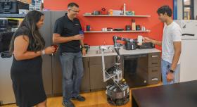 A CoBot MoMa robot hands a student a mug in a simulated apartment at UT Austin while a professor and a fellow student look on and smile.