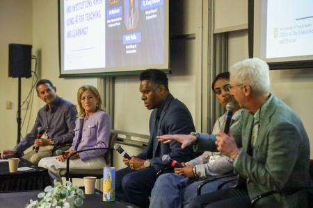 Five individuals are seated on chairs in front of an audience during a panel discussion. Each panelist holds a microphone.
