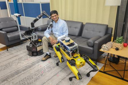 Joydeep Biswas, an associate professor of computer science who will lead the team awarded the NSF grant, sits on a couch smiling, flanked by two robots. On his right, a service robot with a cylindrical base lays its mechanical arm on Biswas' shoulder; to Biswas' left is a robot dog facing him, standing on all four legs.